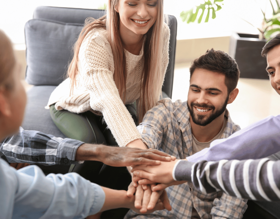 Fünf Personen sitzen gemeinsam in einem Wohnzimmer und legen ihre Hände übereinander als Zeichen von Teamgeist und Zusammenhalt.