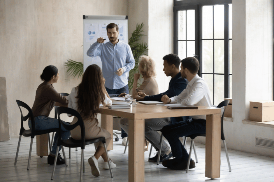Sechs Personen in einem modernen Büro bei einer Besprechung, eine Person präsentiert an einem Flipchart vor großen Fenstern.